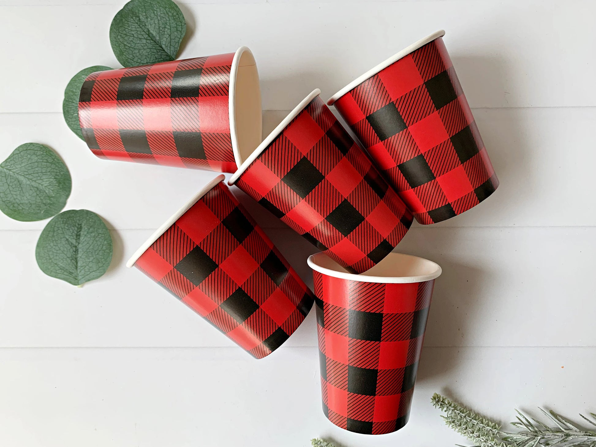 Five red and black buffalo plaid paper cups arranged on a white wood background with decorative green leaves and pine branches.