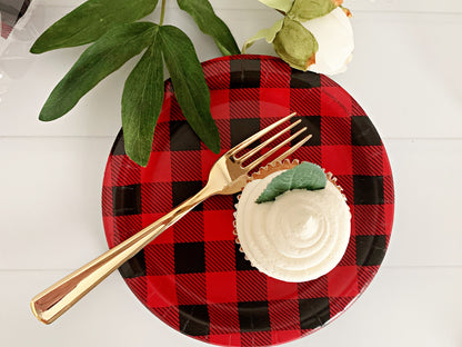 A top-down view of a red and black buffalo plaid paper plate on a white wooden surface, styled with a gold metal fork and a white frosted cupcake with a green leaf decoration.