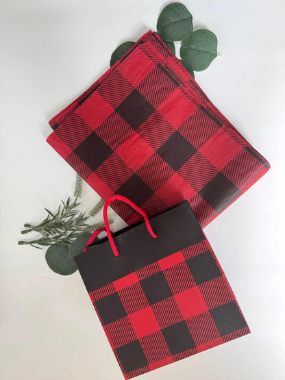 Red and black buffalo plaid gift bags and matching tissue paper with green eucalyptus leaves, arranged on a clean white background.