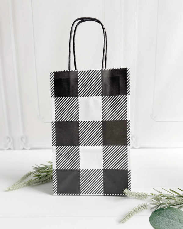 A black and white buffalo plaid paper gift bag with black twisted handles, standing upright on a white surface with decorative greenery in the foreground for wrapping Christmas gifts and baby shower favors