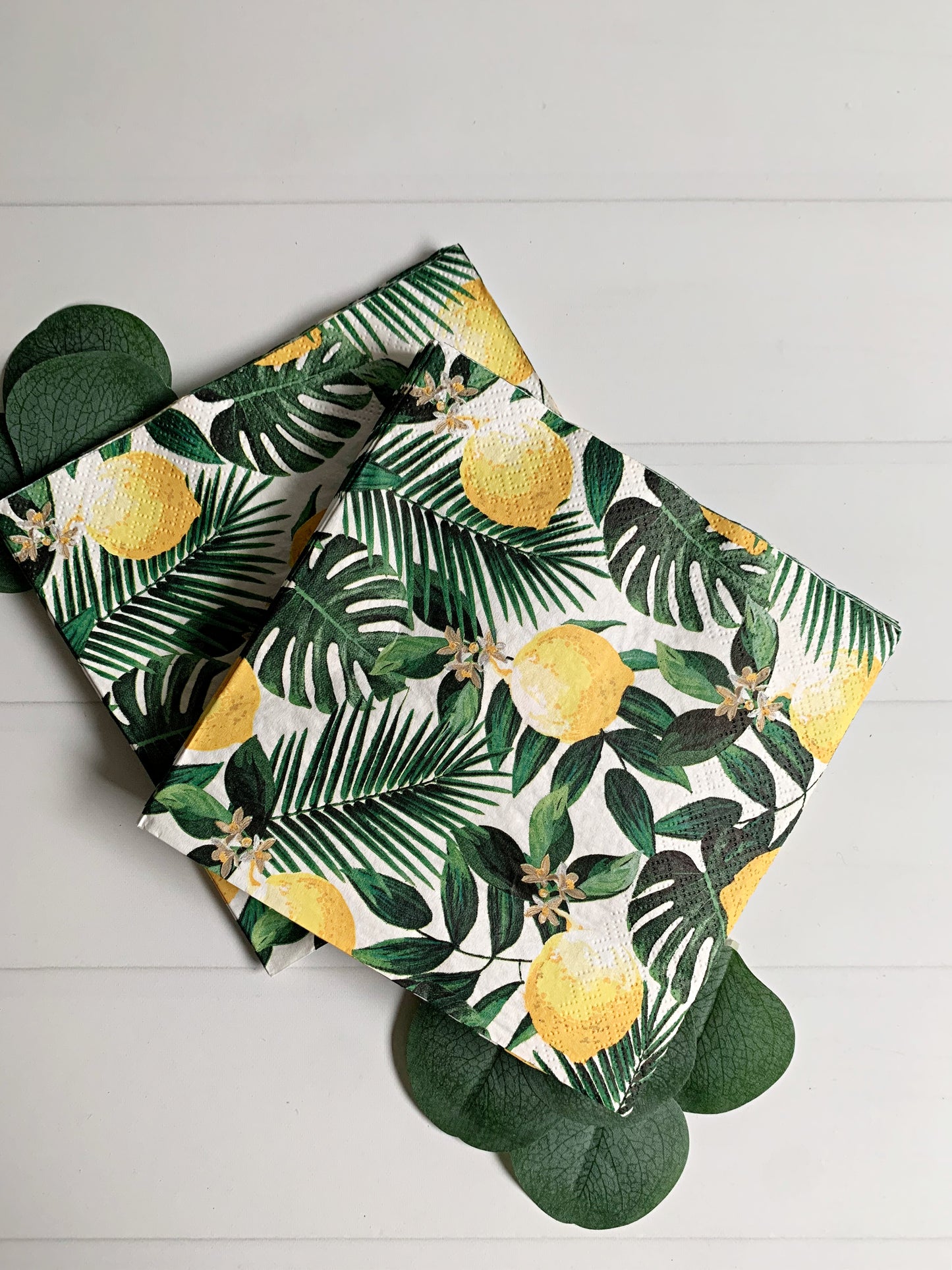A folded elegant luncheon napkins featuring a tropical print of yellow lemons, palm leaves, and monstera leaves on a white background. It is styled on a white wood plank surface with green scalloped paper cutouts underneath.