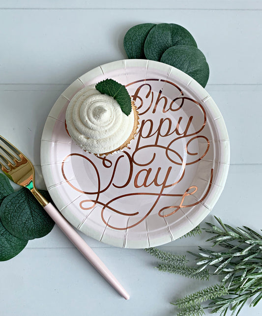 Paper birthday plate with rose gold ‘Happy Day' text, cupcake, and green leaves on a light background