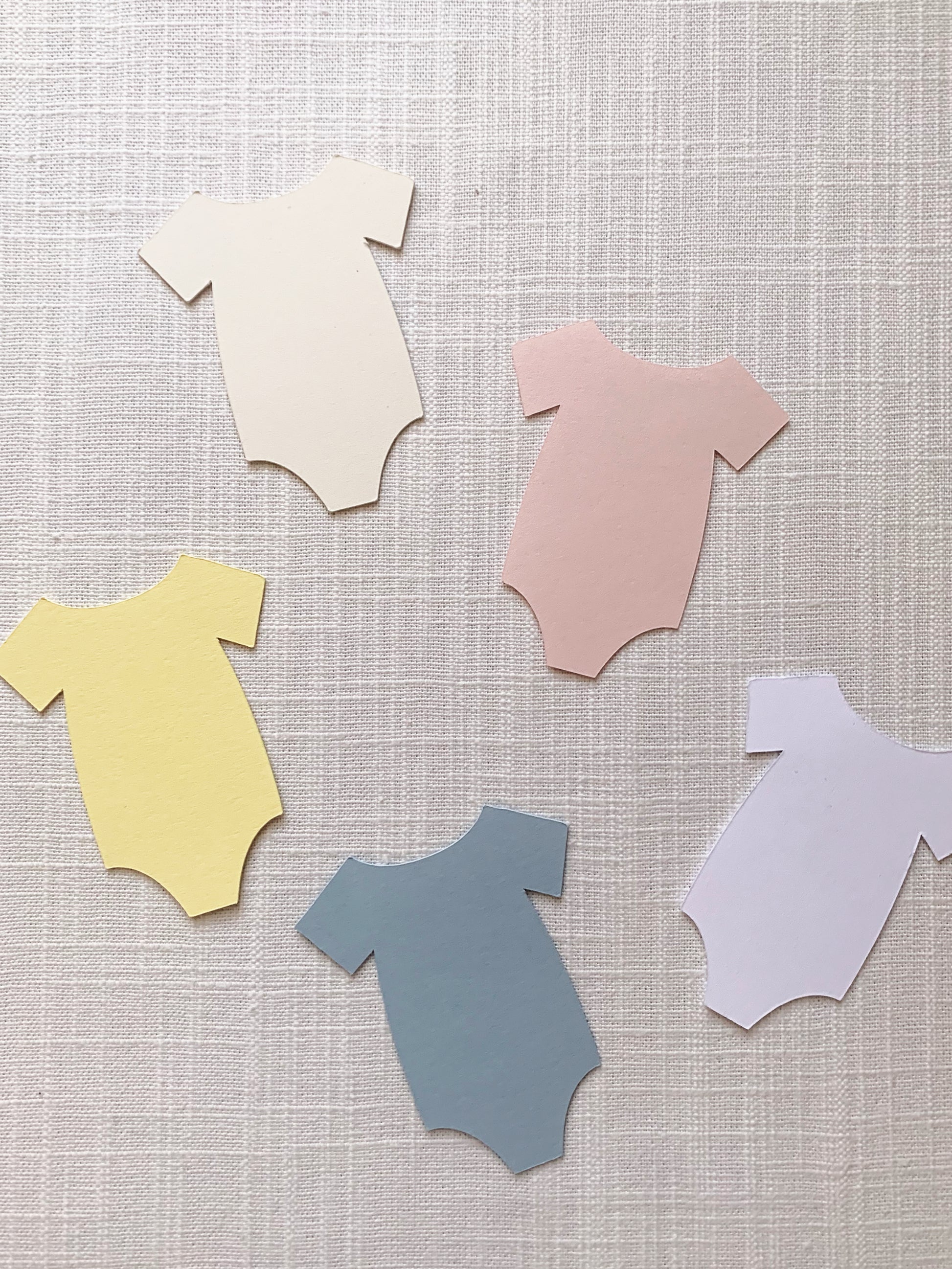 Five paper cutout baby bodysuit shapes in cream, blush pink, yellow, slate blue, and white, arranged on a light beige textured fabric background.