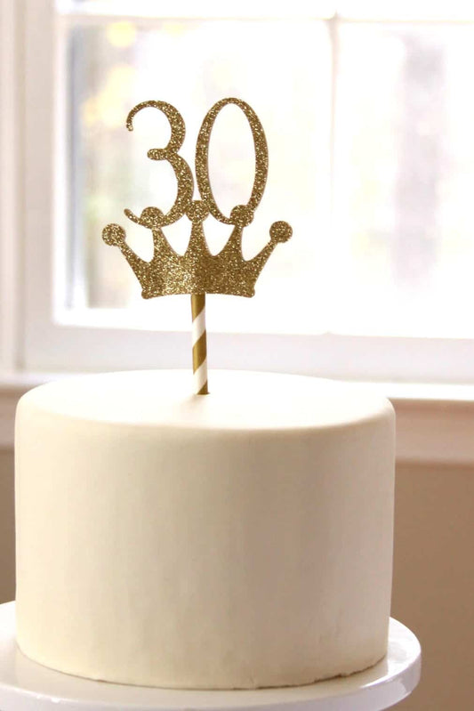 White cake with a gold '30' and crown topper on a blurred background