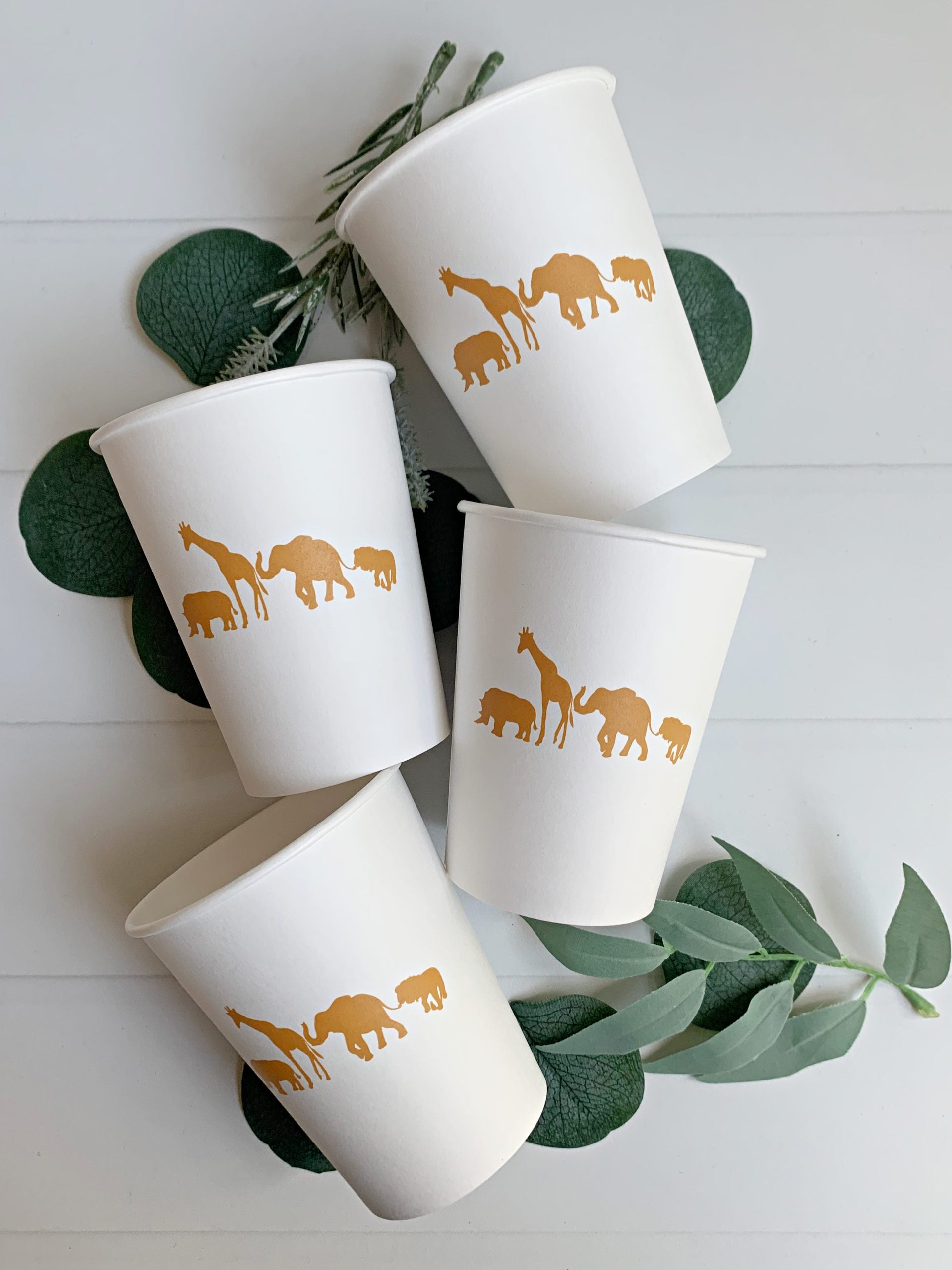 Four white paper cups with gold safari animal prints on a white surface with green leaves.