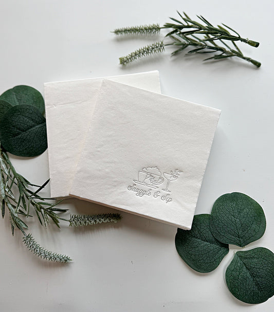 White napkin with 'Snuggle and Sip' logo surrounded by green leaves on a white background