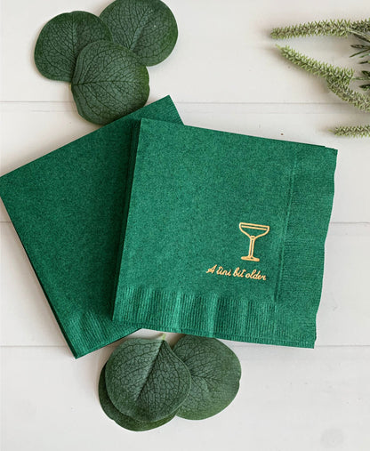 Green napkins with gold text on a white surface with green coupe glass an tini bit older in gold foil
